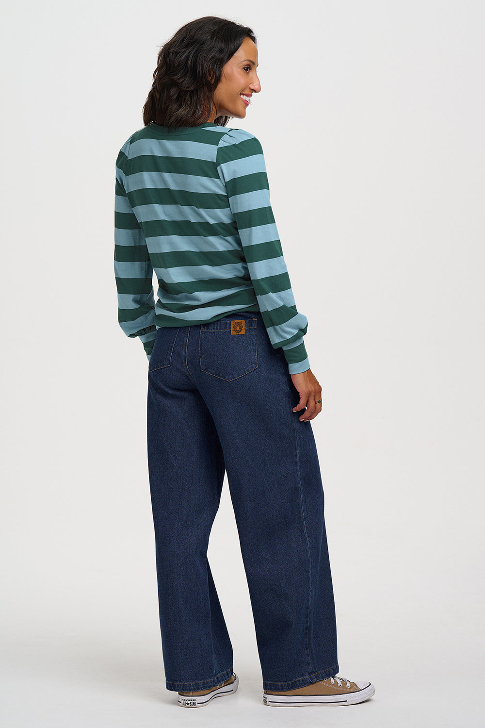 A woman shown from the back wearing a green and light blue striped long-sleeve shirt, dark blue wide-leg jeans, and brown sneakers, against a plain light gray studio background.