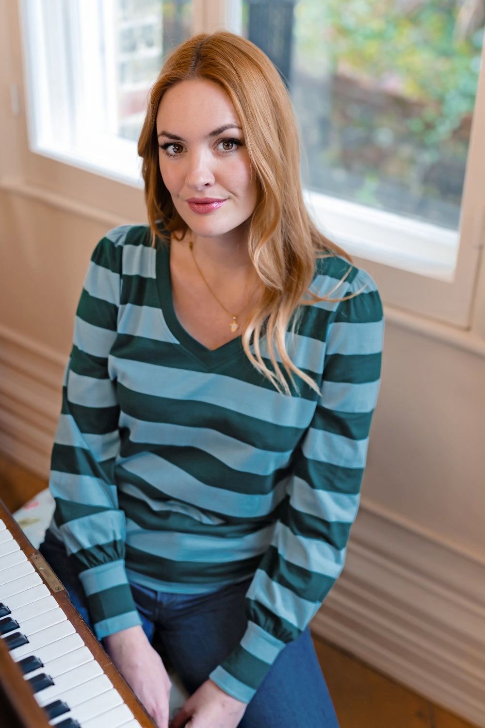 A woman with blonde hair sitting indoors near a piano, wearing a long-sleeve V-neck top with horizontal stripes in dark green and light blue.