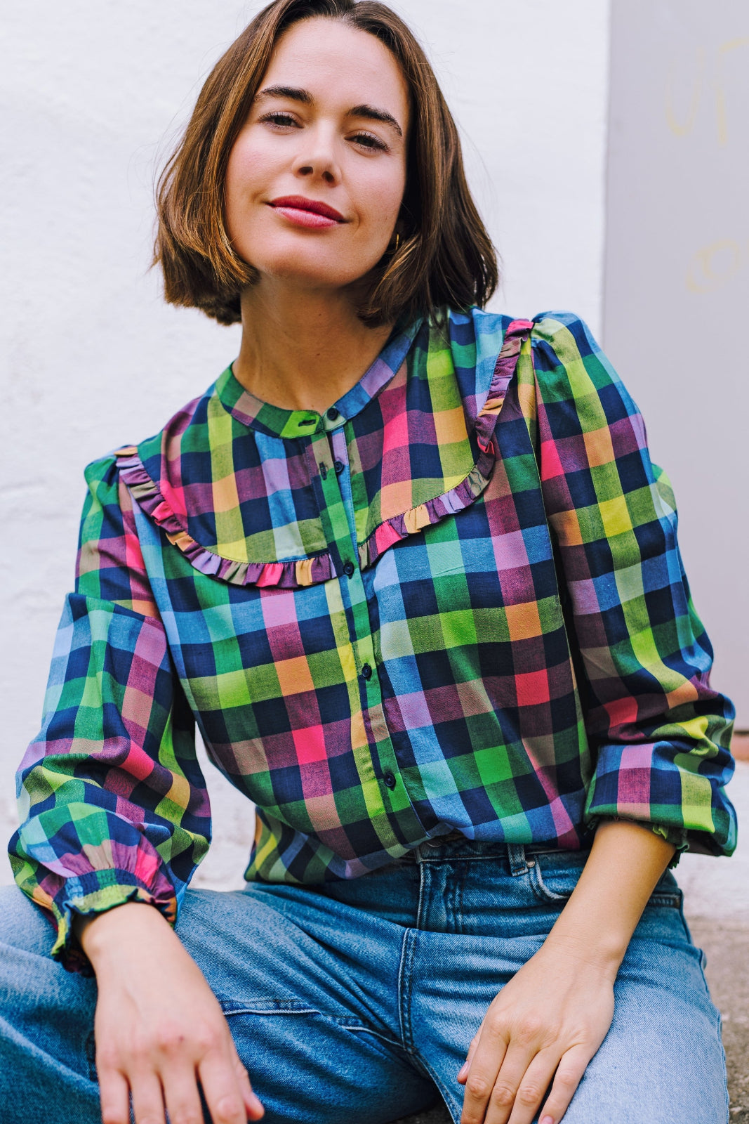 A woman wearing a multicolored checkered shirt with a grandad collar and frill details on the chest and sleeves, paired with blue jeans.
