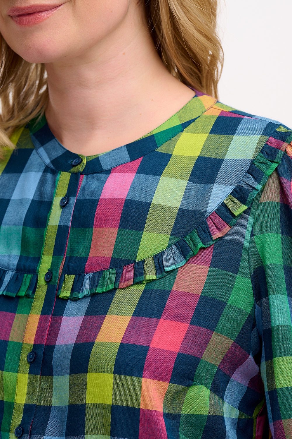 A woman wearing a multicolored checkered shirt with a grandad collar and frill details on the chest and sleeves, paired with blue jeans.