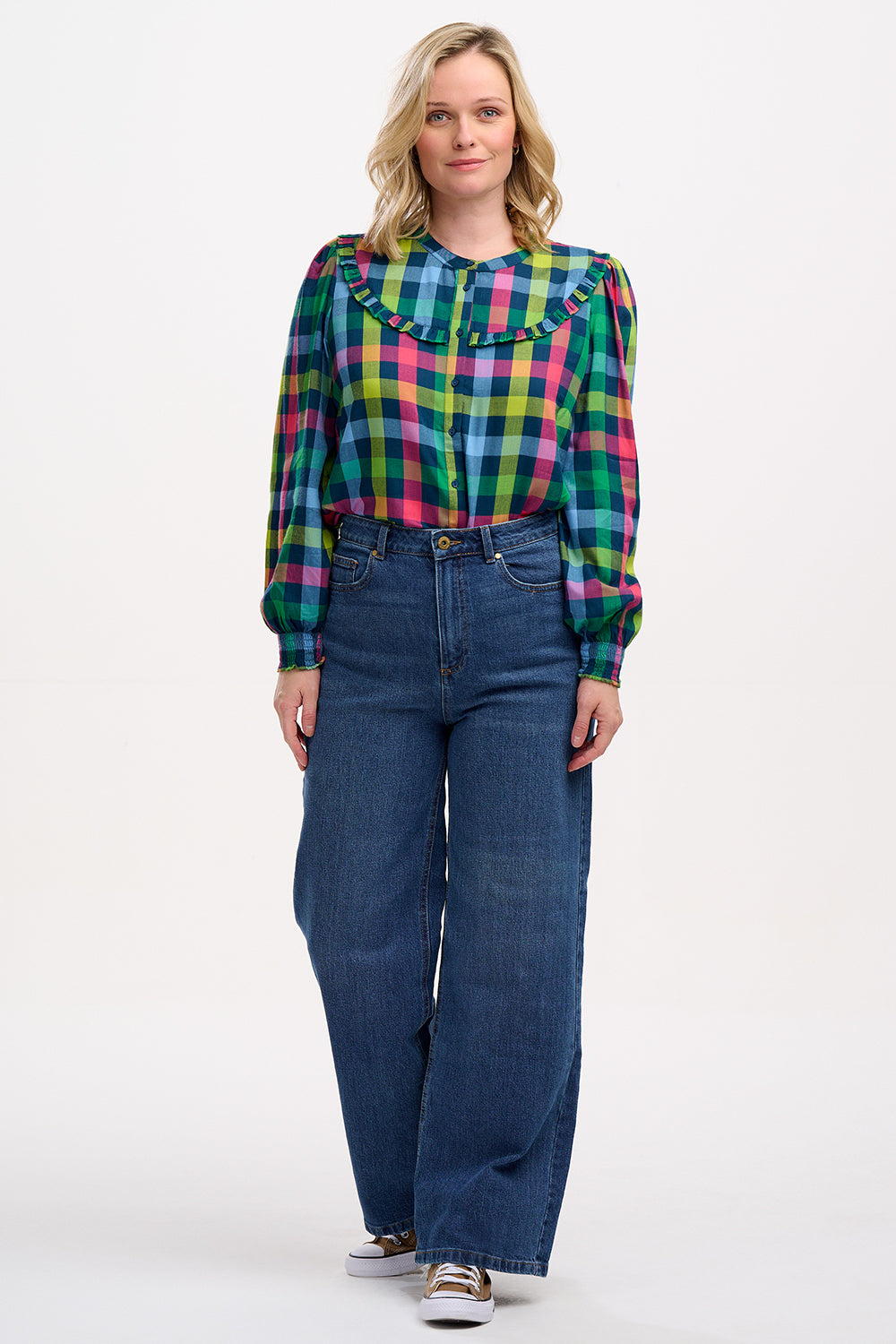 A woman wearing a multicolored checkered shirt with a grandad collar and frill details on the chest and sleeves, paired with blue jeans.