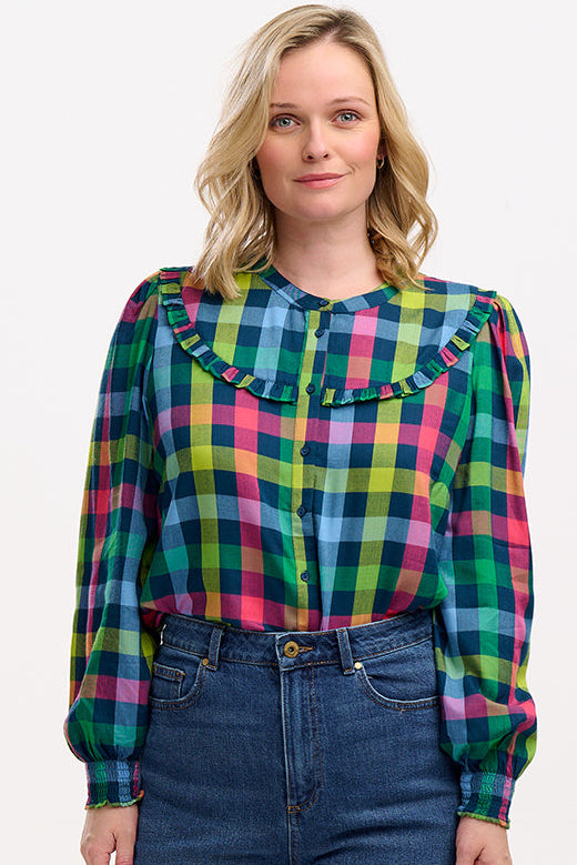 A woman wearing a multicolored checkered shirt with a grandad collar and frill details on the chest and sleeves, paired with blue jeans.