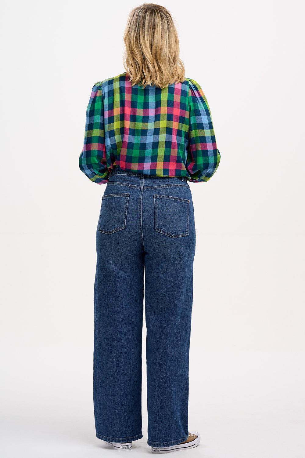 A woman wearing a multicolored checkered shirt with a grandad collar and frill details on the chest and sleeves, paired with blue jeans.