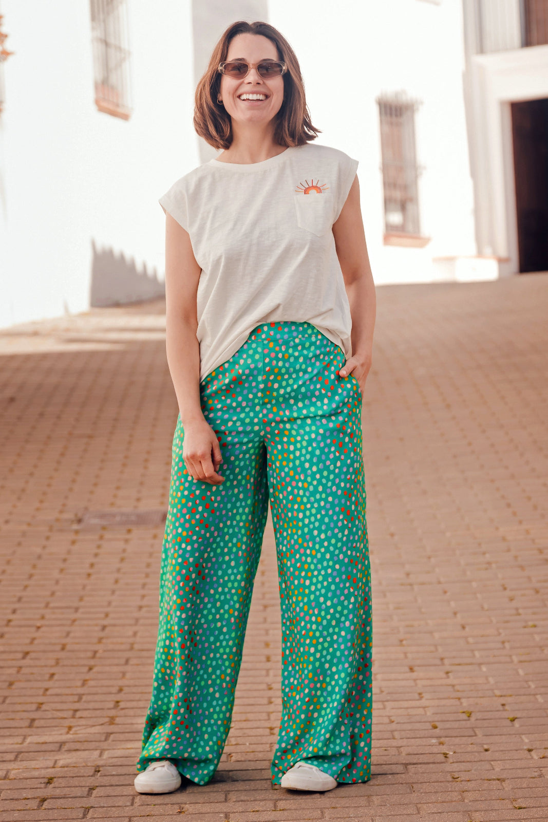 Woman wearing a white t-shirt and green patterned pants standing on a brick pavement.