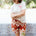 Woman wearing a white t-shirt with a palm tree design and red patterned shorts, standing in a garden.
