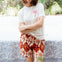 Woman wearing a white t-shirt with a palm tree design and red patterned shorts, standing in a garden.
