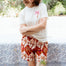 Woman wearing a white t-shirt with a palm tree design and red patterned shorts, standing in a garden.