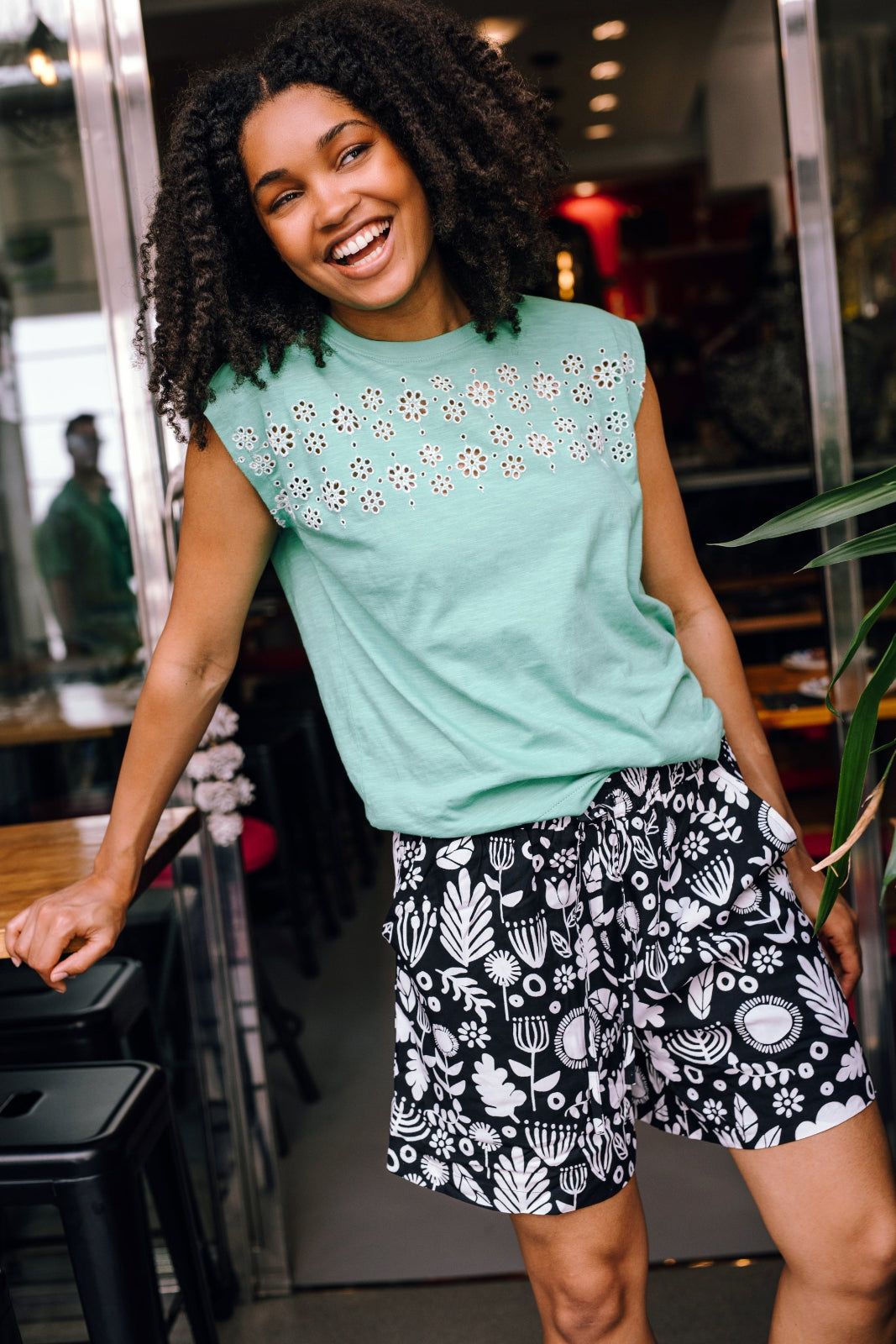 A person wearing a green sleeveless top with a floral folk print and black shorts with a similar floral pattern.