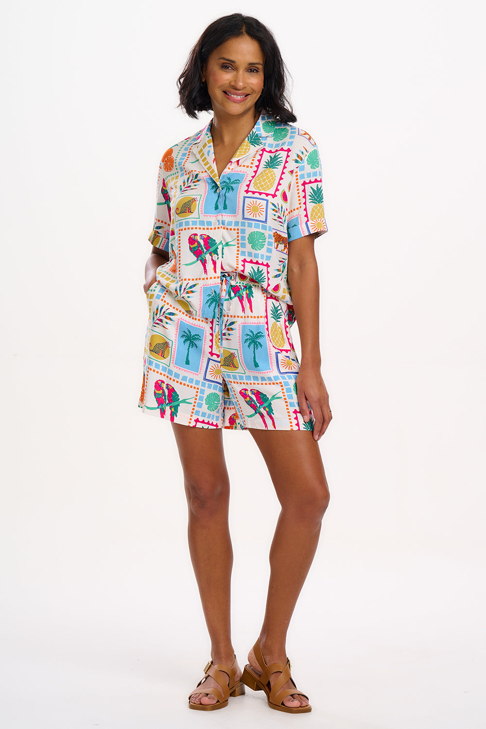 A woman standing, wearing off-white shorts with a multicolor jungle tile print, paired with a matching top, and brown sandals.