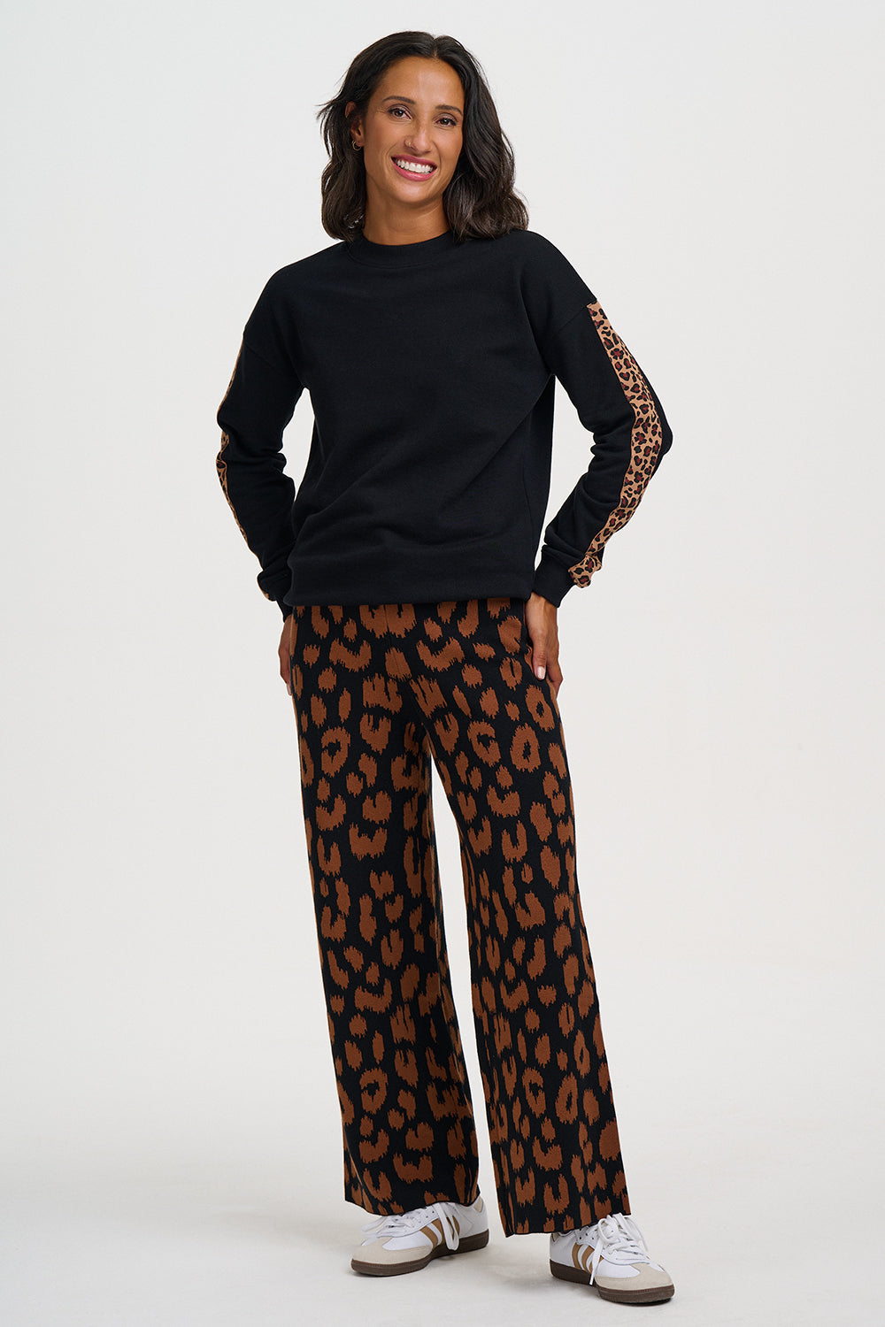 A woman modeling a black crewneck sweatshirt with leopard-print patterned sleeves and matching wide-leg pants in a brown and black leopard print, paired with white sneakers, against a light gray studio background.
