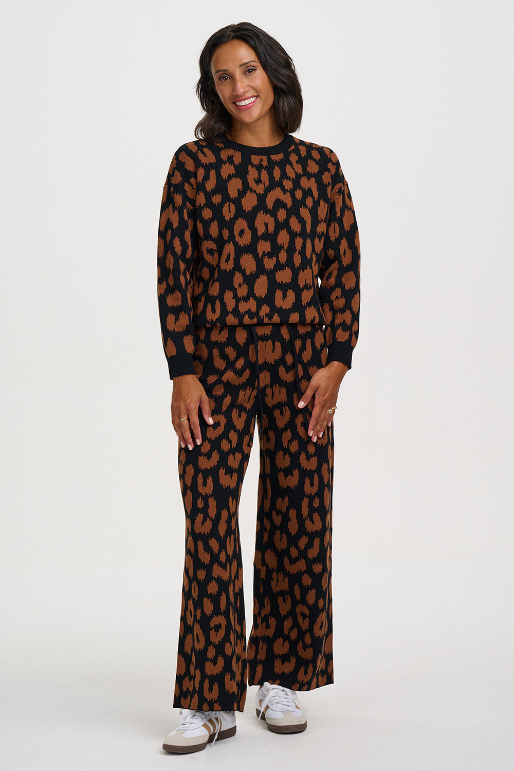 A woman modeling a matching two-piece lounge set featuring a brown and black leopard print pattern, consisting of a long-sleeve top and wide-leg pants, paired with white sneakers against a plain light gray background.