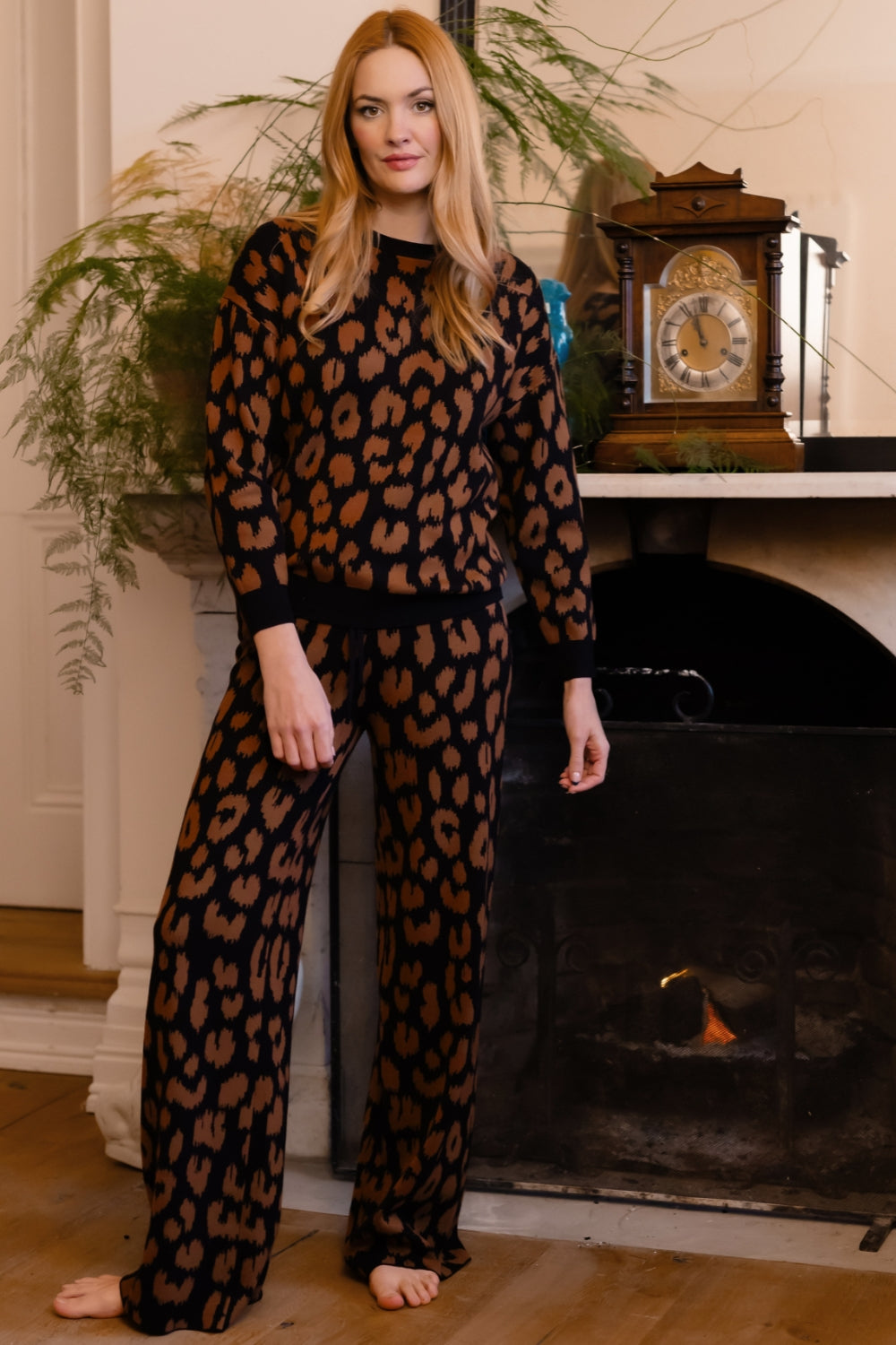 A woman stands barefoot in front of a fireplace wearing a matching long-sleeve top and wide-leg pants set in a black and brown leopard print fabric.
