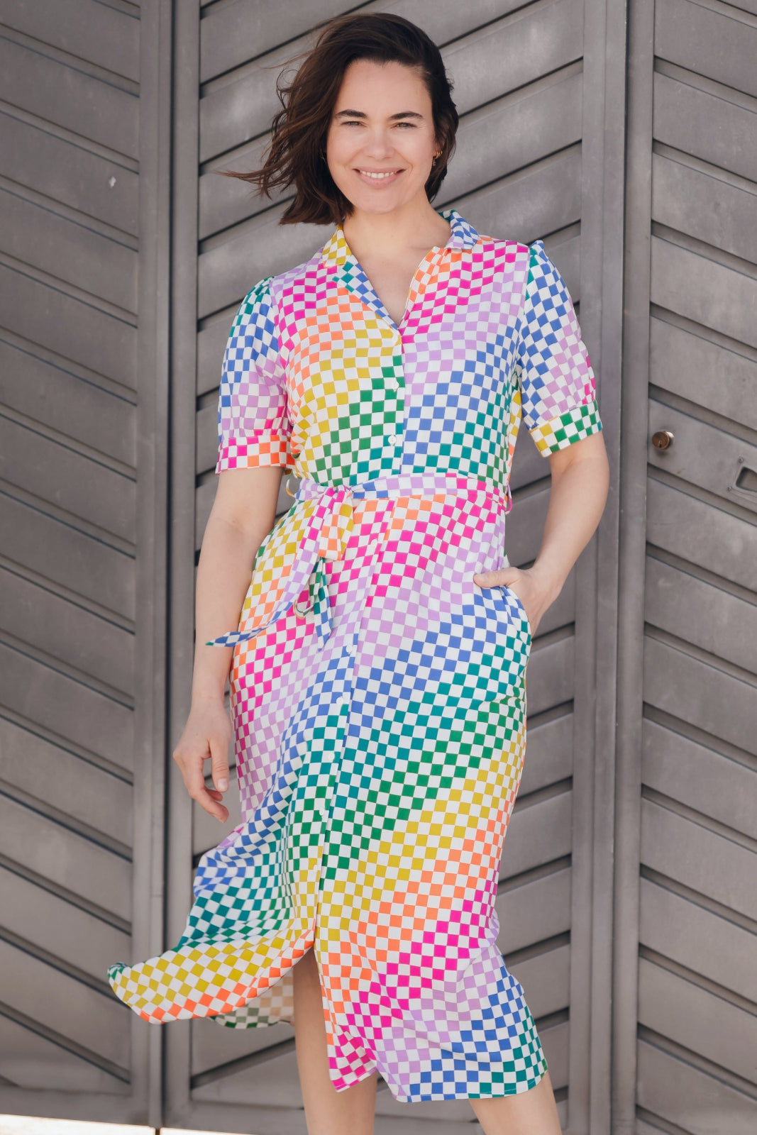 A model wearing a midi length shirt dress with a rainbow checkerboard pattern, featuring a lapel collar and puff sleeves.