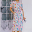 A model wearing an off-white midi-length shirt dress with a Jungle Tile print, featuring colorful tropical wildlife, fruit, and plants. The dress has a button placket, puff sleeves, and a tie waist.
