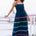 A model wearing a navy sundress with rainbow-colored Ric Rac embellishments on the hem and shoulder straps, standing by a railing.