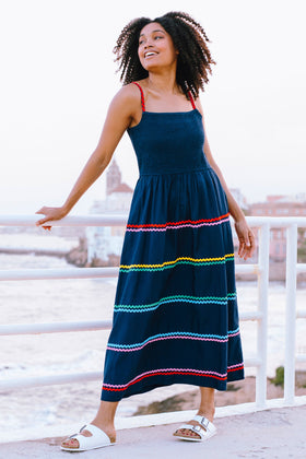 A model wearing a navy sundress with rainbow-colored Ric Rac embellishments on the hem and shoulder straps, standing by a railing.
