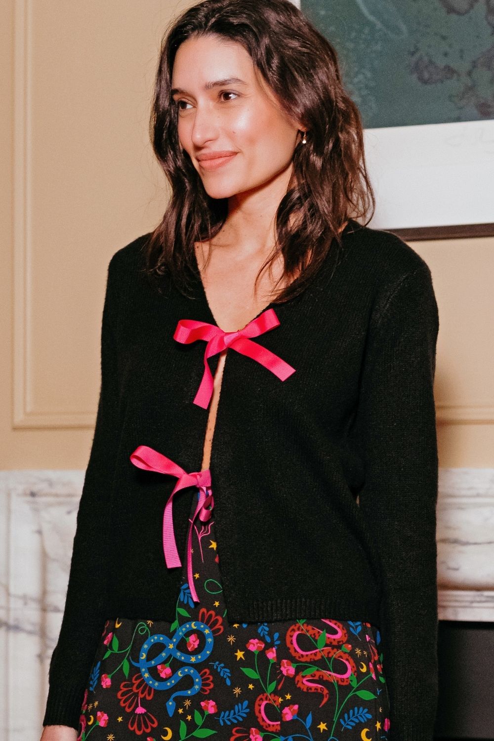 Woman wearing a black cardigan with pink bows and an embroidered skirt indoors.