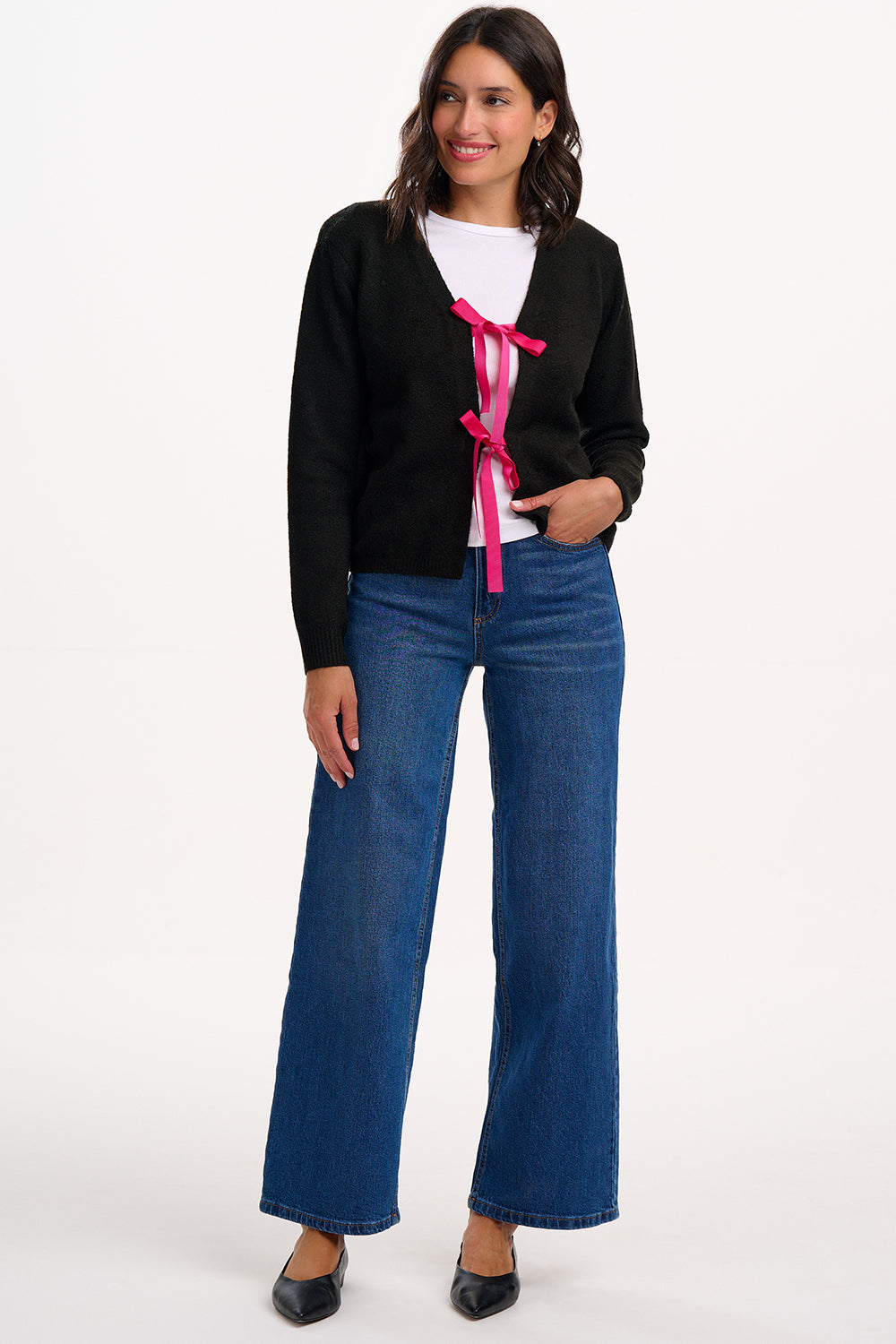 Woman wearing a black cardigan with pink tie, white shirt, and blue jeans on a white background