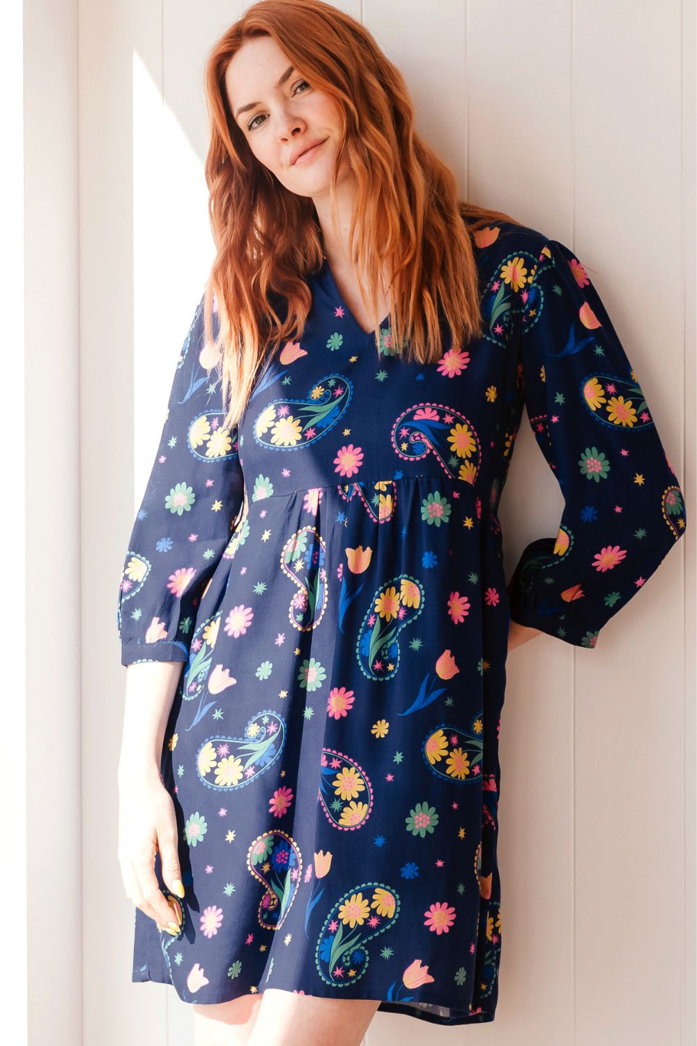 Woman wearing a colorful dress with a floral pattern against a white wall.