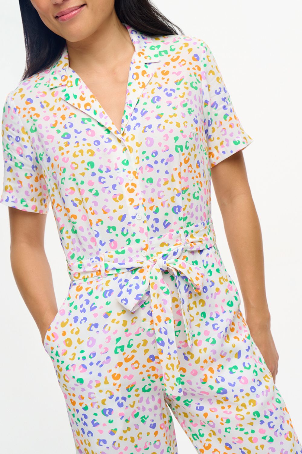 A model wearing an off-white jumpsuit with a mottled leopard print design, featuring short sleeves, a revere collar, side pockets, and a tie waist.