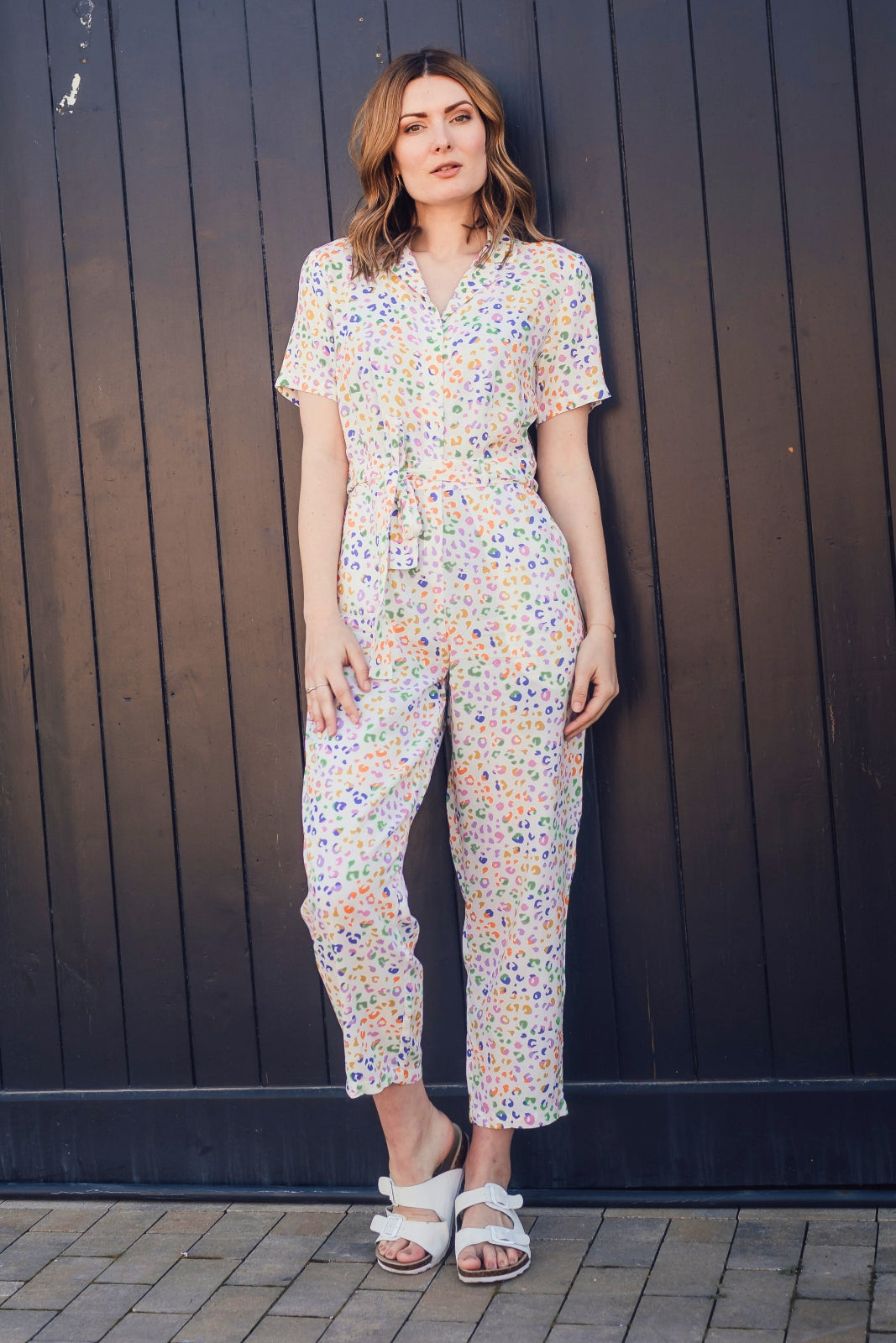 Woman wearing a floral jumpsuit standing against a dark wooden wall.