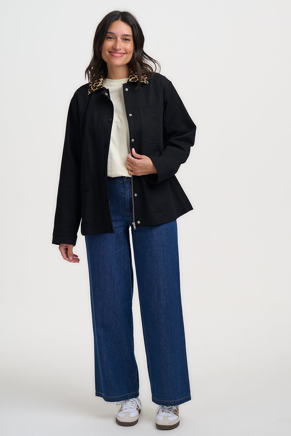 Woman wearing a black jacket, white shirt, blue jeans, and white sneakers on a white background
