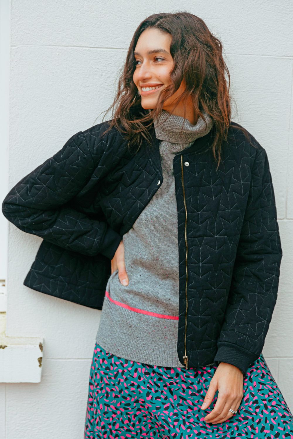 Woman wearing a black quilted jacket, gray sweater, and patterned skirt against a white wall.