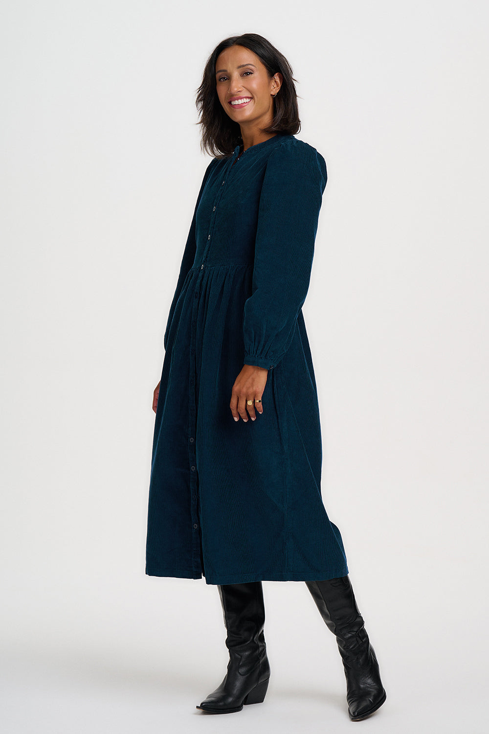 A woman modeling a long-sleeved navy blue midi dress with a gathered waist and black knee-high leather boots, photographed against a plain light gray studio background.