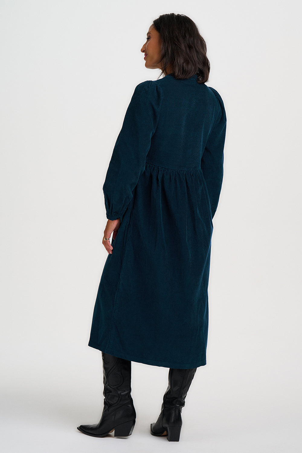 A woman shown from the back wearing a dark teal corduroy midi dress with long sleeves and black knee-high boots, photographed against a plain light gray studio background.