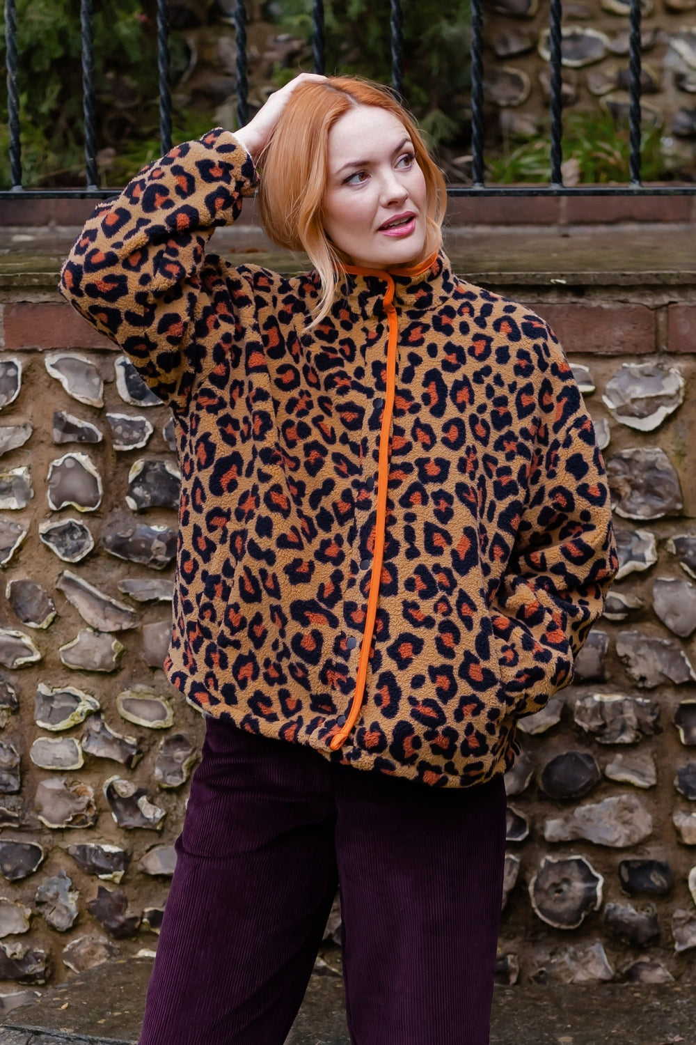 A woman with red hair wearing a leopard print fleece jacket with an orange zipper, paired with dark purple pants, standing in front of a stone wall.