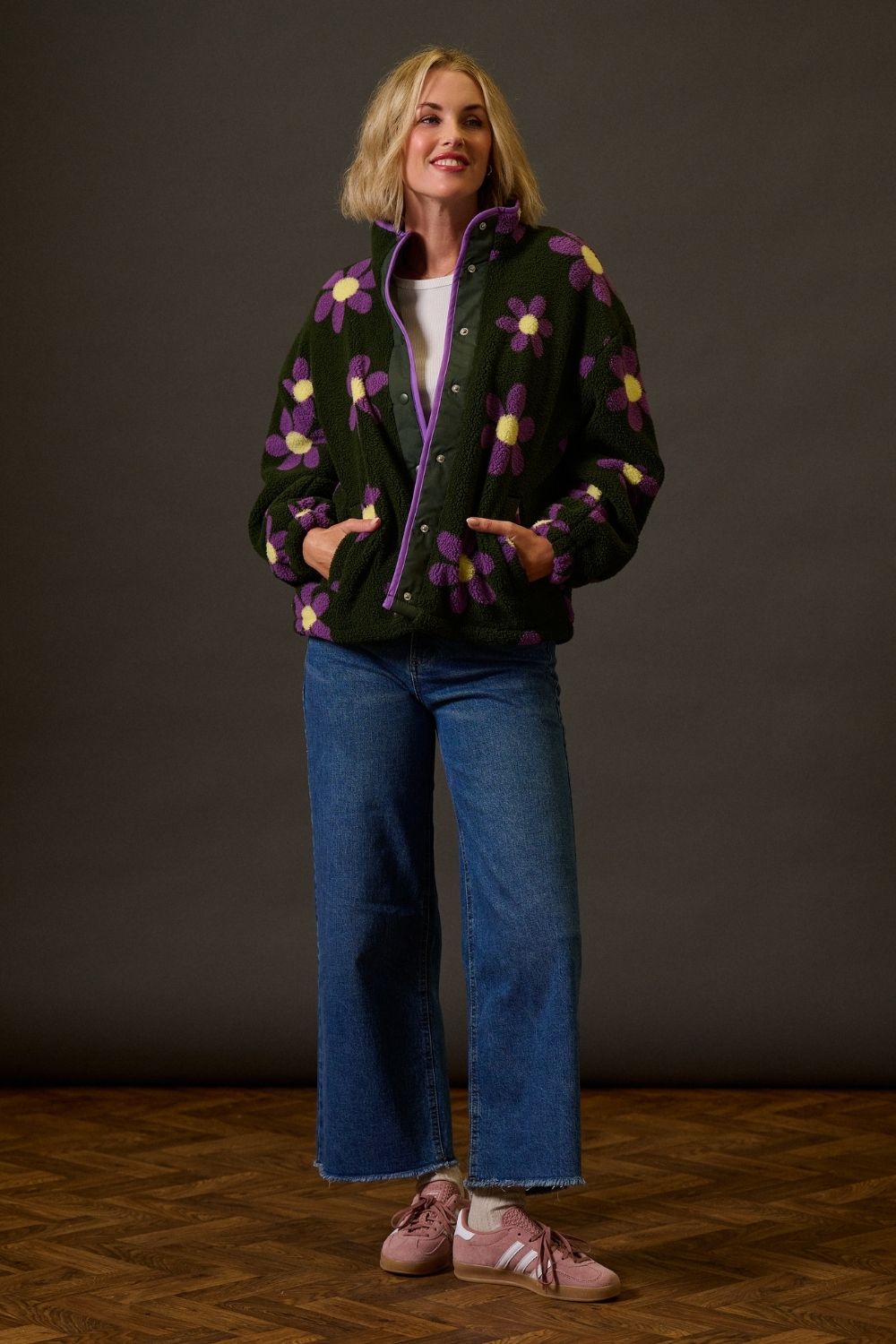 Woman wearing a dark jacket with colorful floral patterns, blue jeans, and pink shoes on a wooden floor.