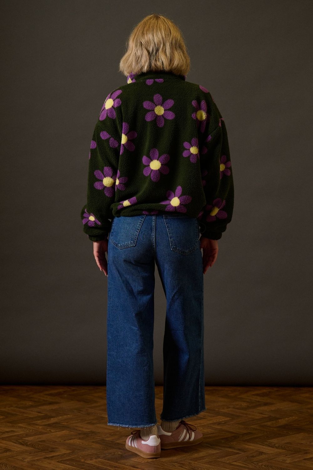 Person wearing a dark jacket with floral patterns and blue jeans against a brown background