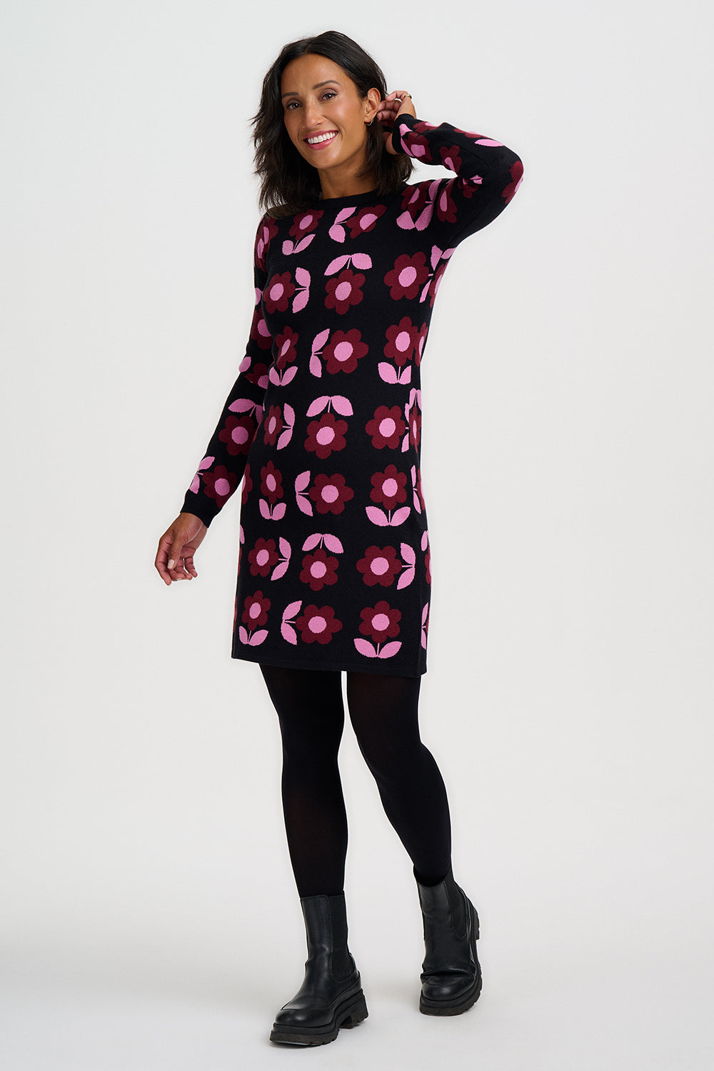 A woman modeling a long-sleeve black knit dress with a pink and magenta floral pattern, paired with black tights and black ankle boots, against a light gray studio background.