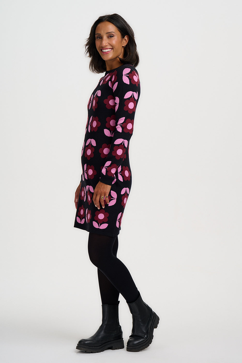 A woman modeling a long-sleeve black knit dress with a pink and magenta floral pattern, styled with black tights and black ankle boots against a light gray studio background.