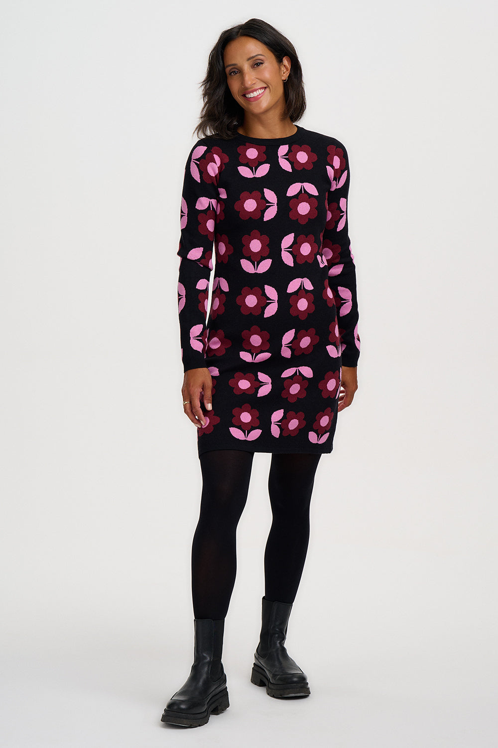 A woman modeling a black long-sleeve knit dress with a pink and magenta floral pattern, paired with black tights and black boots, against a plain white studio background.