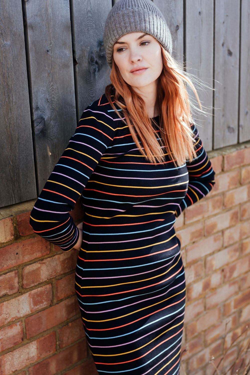 Woman wearing a striped dress and beanie against a brick wall.