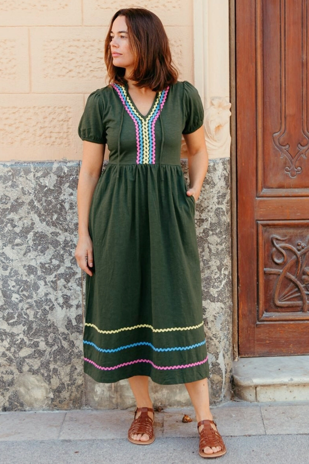 Woman wearing a green dress with colorful patterns standing against a textured wall.