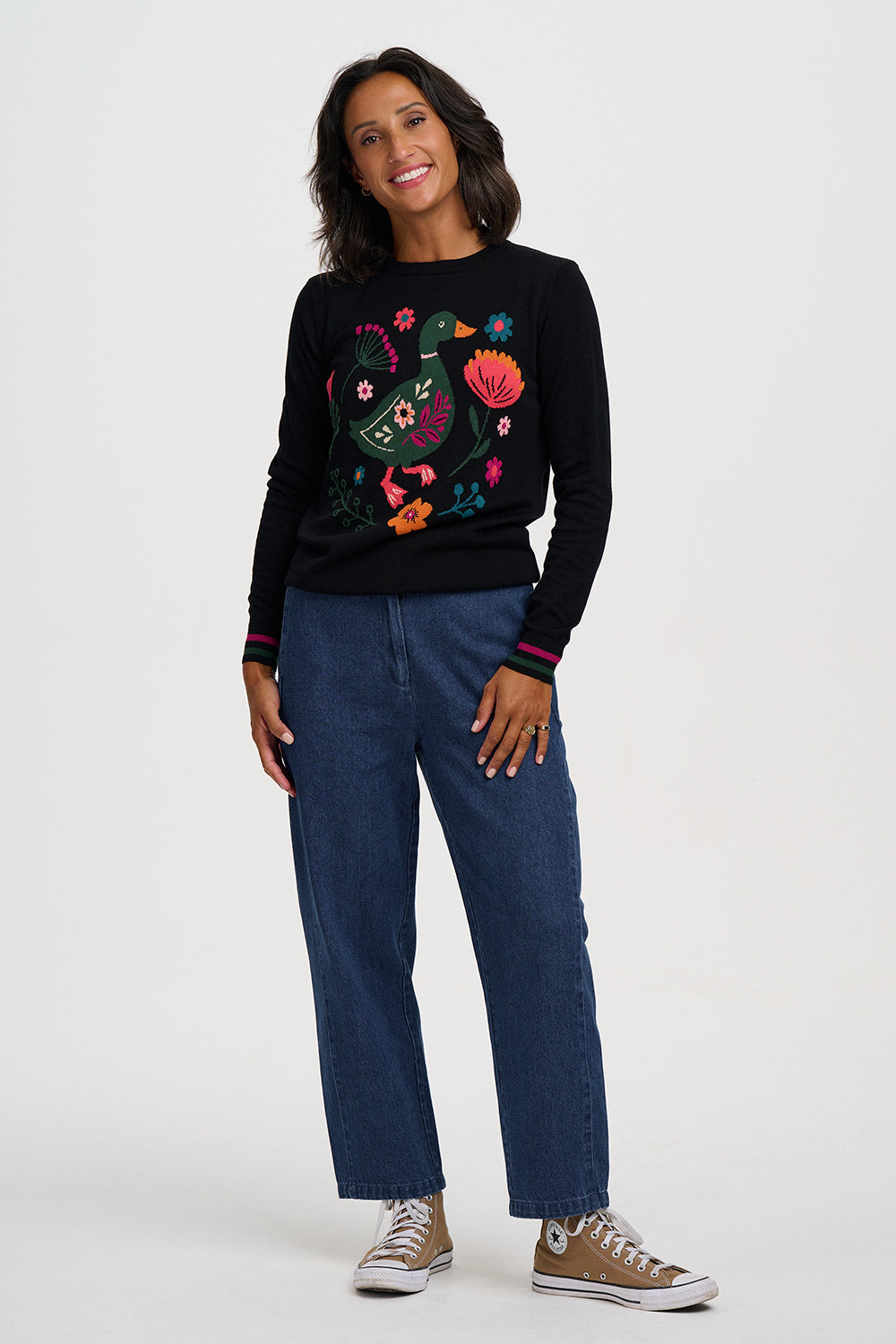 A woman modeling a black long-sleeve crewneck sweater with colorful floral embroidery on the front, paired with blue jeans and brown sneakers, against a light gray studio background.
