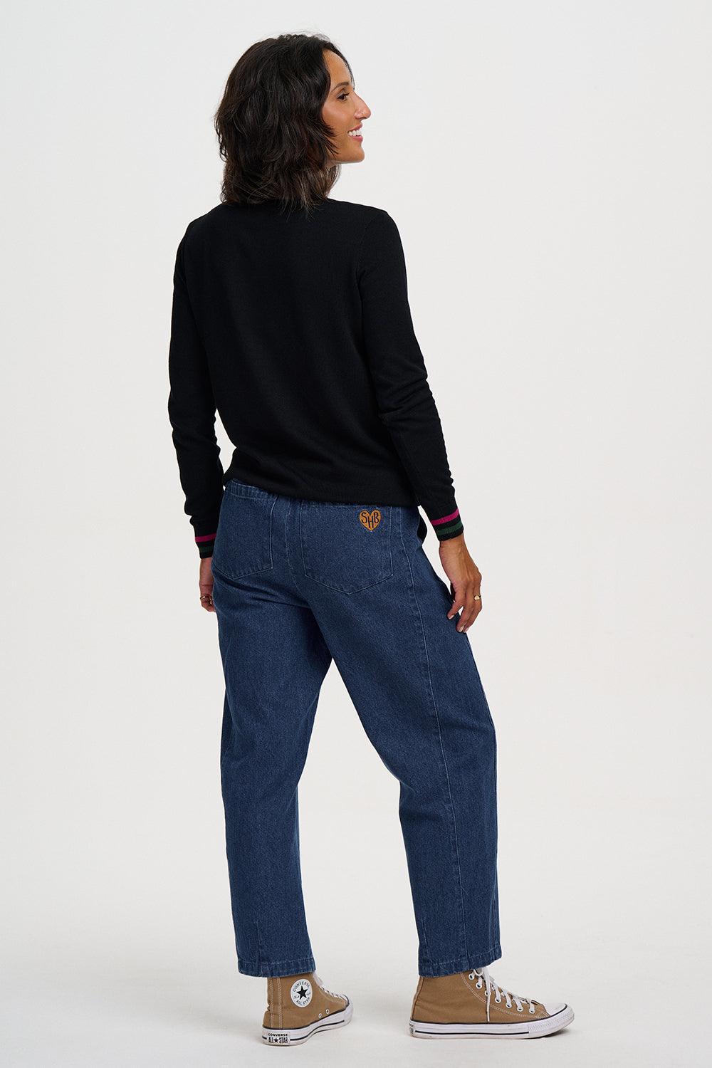 A woman is shown from the back wearing a black long-sleeve top, blue jeans, and tan sneakers against a plain light gray studio background.