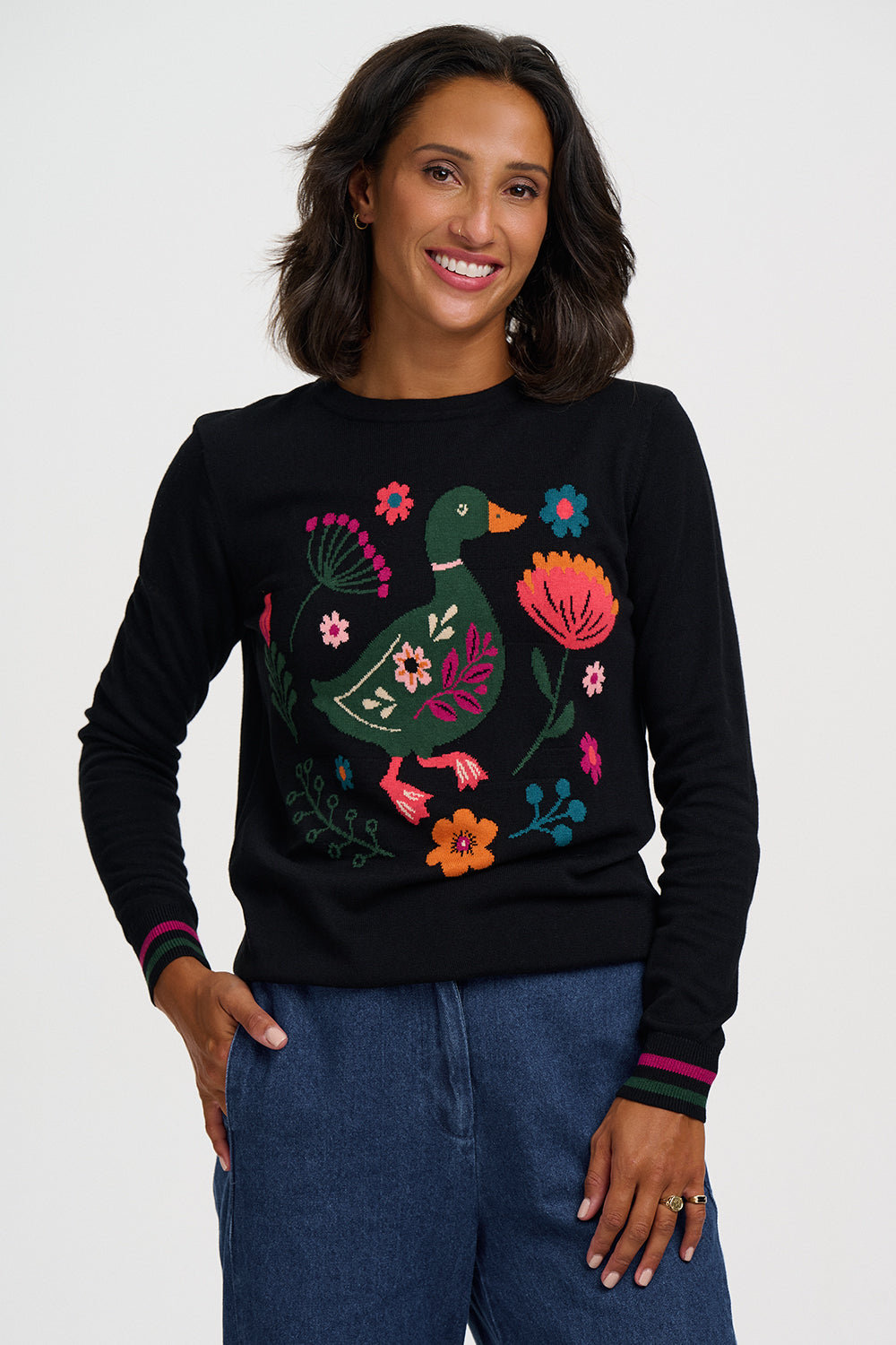 A woman wearing a black crewneck sweater with a colorful embroidered duck and floral design, paired with blue jeans, against a light gray studio background.