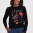 A woman wearing a black crewneck sweater with a colorful embroidered duck and floral design, paired with blue jeans, against a light gray studio background.