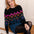A woman with red hair sits on a wooden step in front of a white paneled wall, wearing a black crewneck sweater with a colorful geometric and floral pattern, a denim mini skirt, black tights, and brown leather shoes.