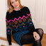 A woman with red hair sits on a wooden step in front of a white paneled wall, wearing a black crewneck sweater with a colorful geometric and floral pattern, a denim mini skirt, black tights, and brown leather shoes.