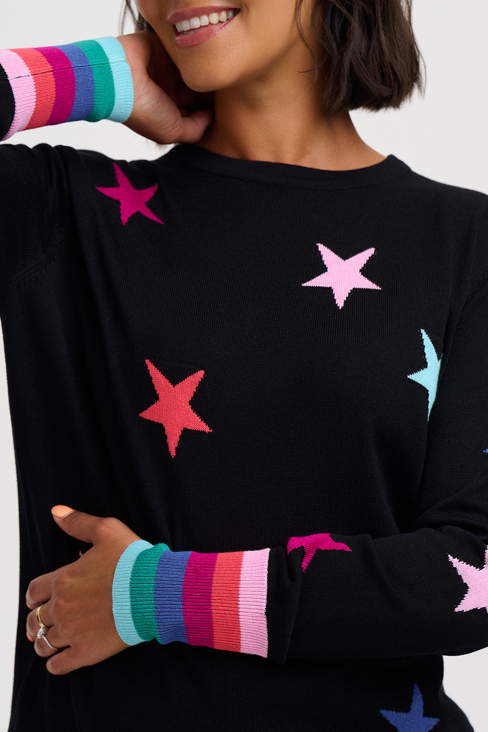 A close-up of a woman wearing a black knit sweater decorated with colorful knitted stars in pink, red, blue, and purple, featuring striped ribbed cuffs in matching rainbow colors.