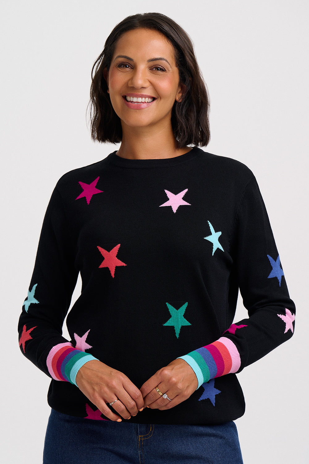 A  woman wearing a black knit sweater decorated with colorful knitted stars in pink, red, blue, and purple, featuring striped ribbed cuffs in matching rainbow colors.