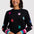 A  woman wearing a black knit sweater decorated with colorful knitted stars in pink, red, blue, and purple, featuring striped ribbed cuffs in matching rainbow colors.