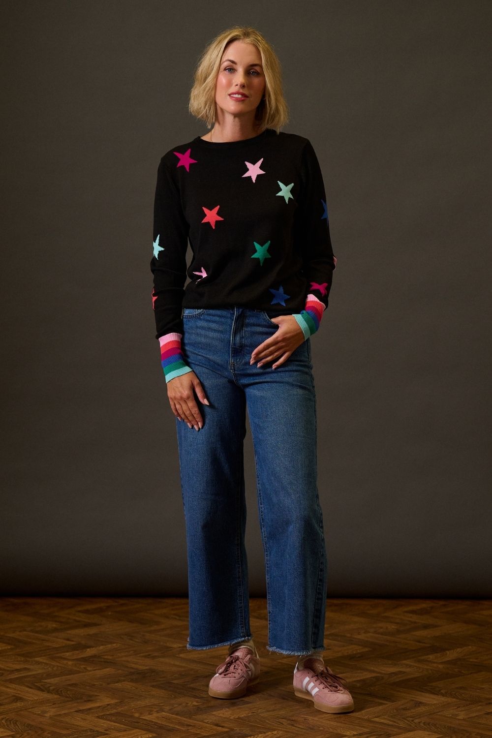 Woman wearing a black sweater with colorful star patterns and blue jeans against a dark background