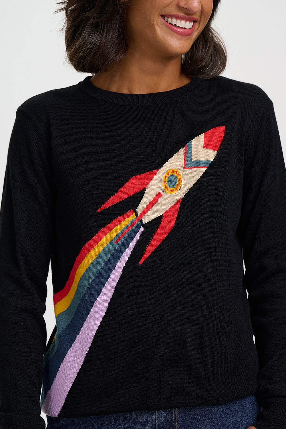 A woman wearing a black crewneck sweater with a colorful knit rocket ship and rainbow design on the front.