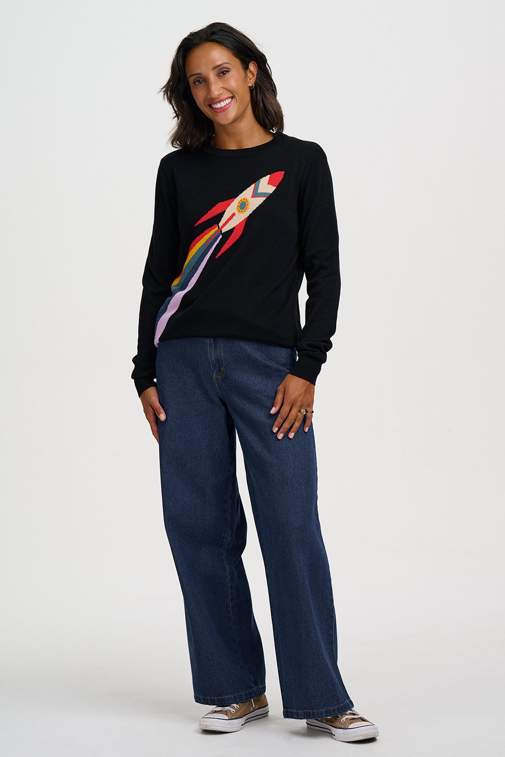 A woman modeling a black crewneck sweater with a colorful rocket ship graphic on the front, paired with dark blue wide-leg jeans and white sneakers, against a light gray studio background.