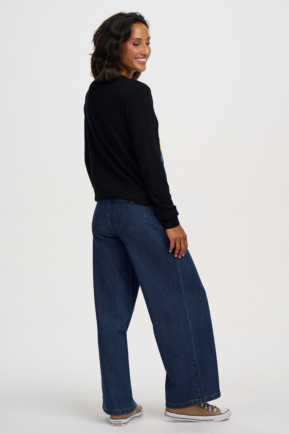 A woman wearing dark blue wide-leg jeans, a black long-sleeve top, and brown sneakers, shown from a side-back angle against a plain light gray studio background.